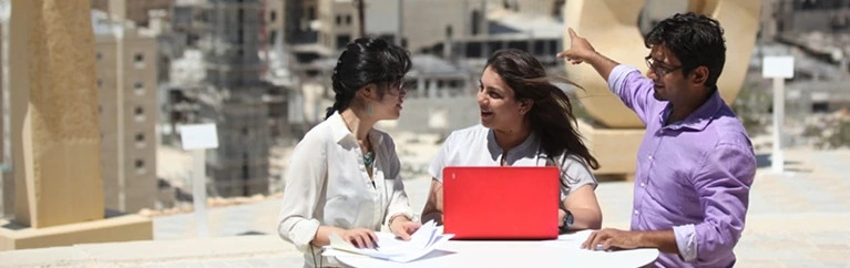 Three students gather around a table with a laptop and papers.